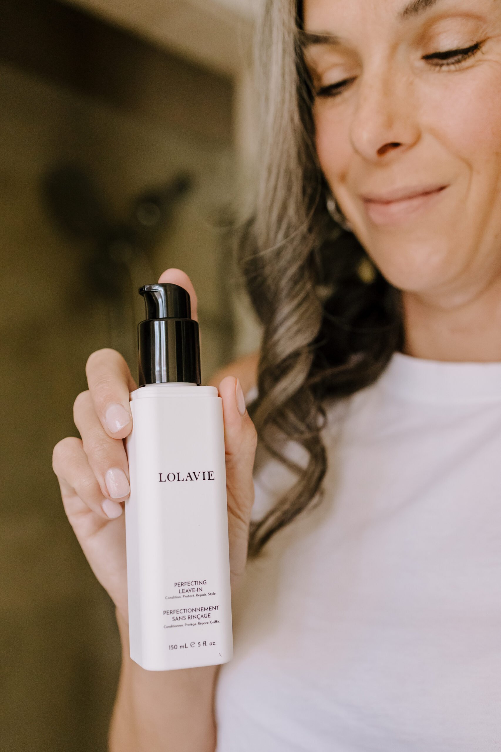 A woman holding up a bottle of LolaVie Perfecting Leave-In.