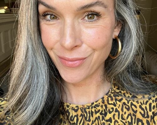 A woman in a yellow leopard print top with long gray hair.