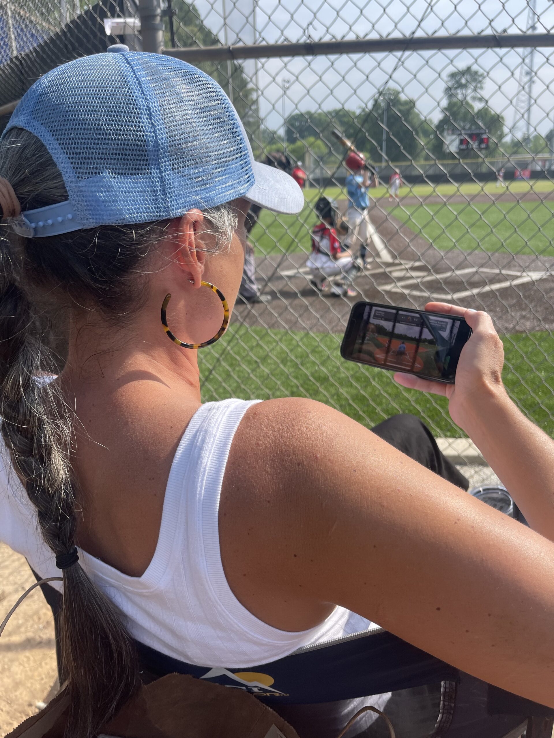 A woman watches a baseball game on her phone while watching another one on the sidelines.
