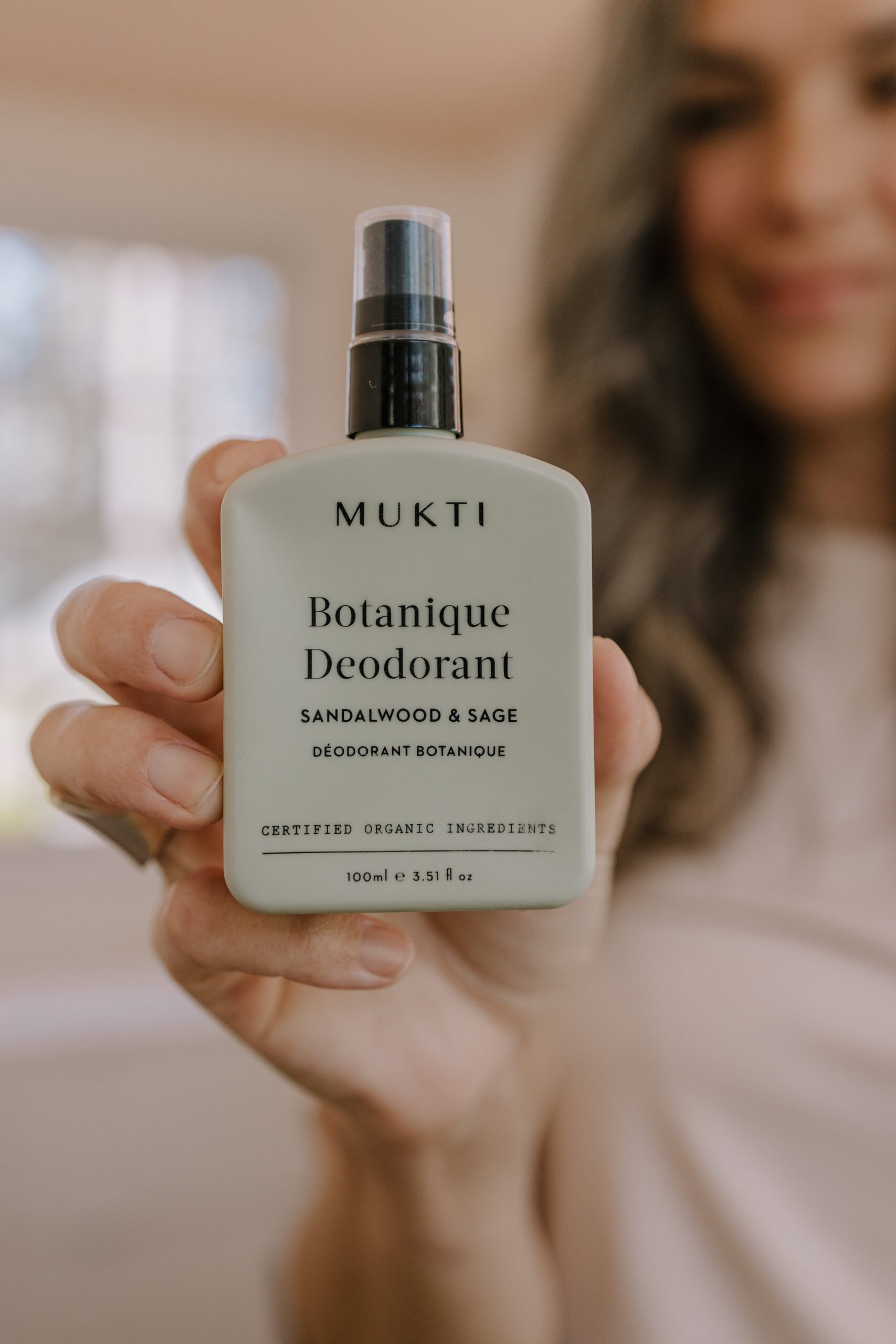A woman holds up a bottle of Mukti Botanique Deodorant. 